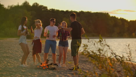 Group-of-five-young-people-are-dancing-in-shorts-and-t-shirts-around-bonfire-in-nature-with-beer.-They-are-clink-and-drink-beer-and-enjoying-the-hot-evening-at-sunset-near-the-lake.